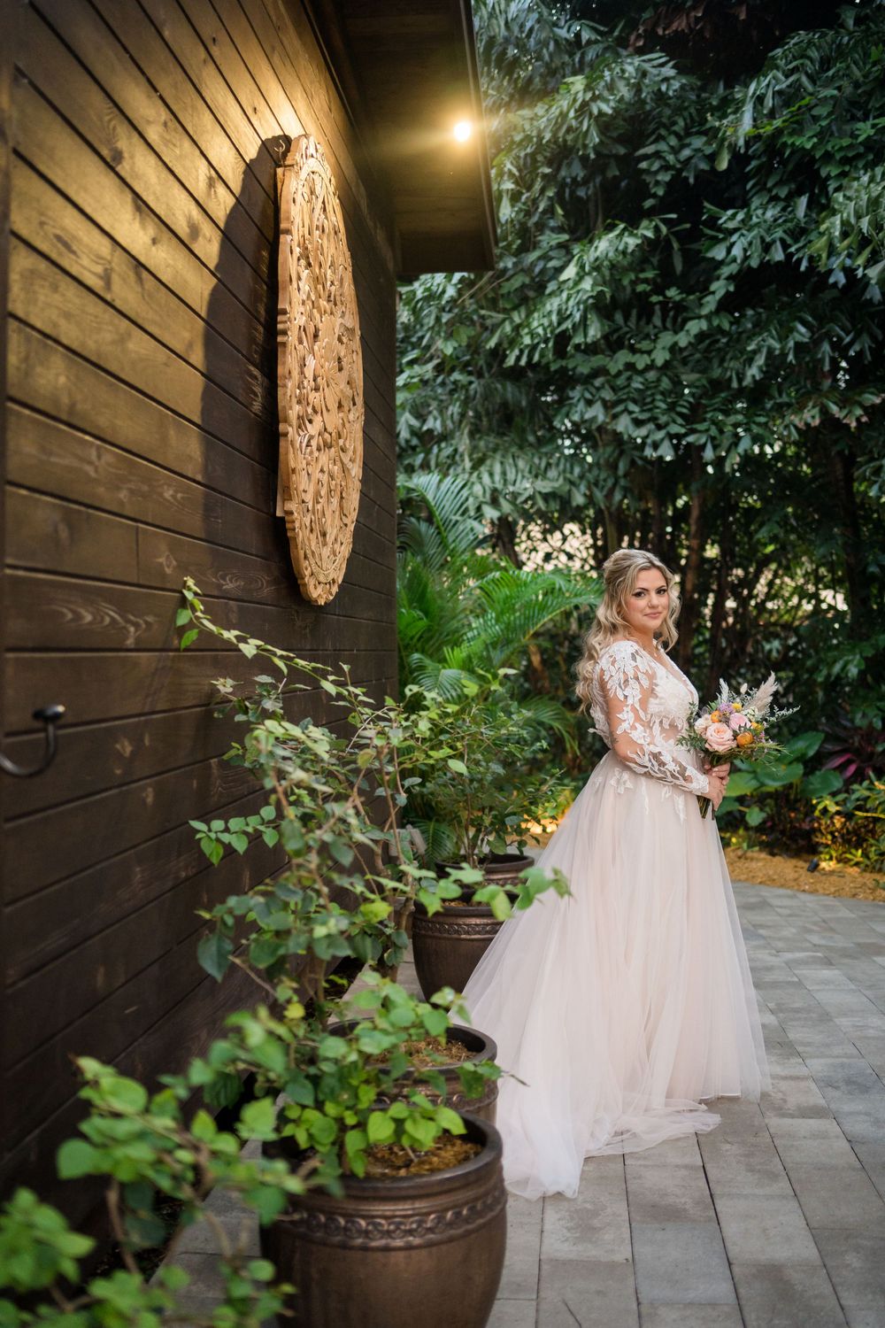 An elegant wedding dress flows beautifully against a wooden wall with mandala art and garden decor.