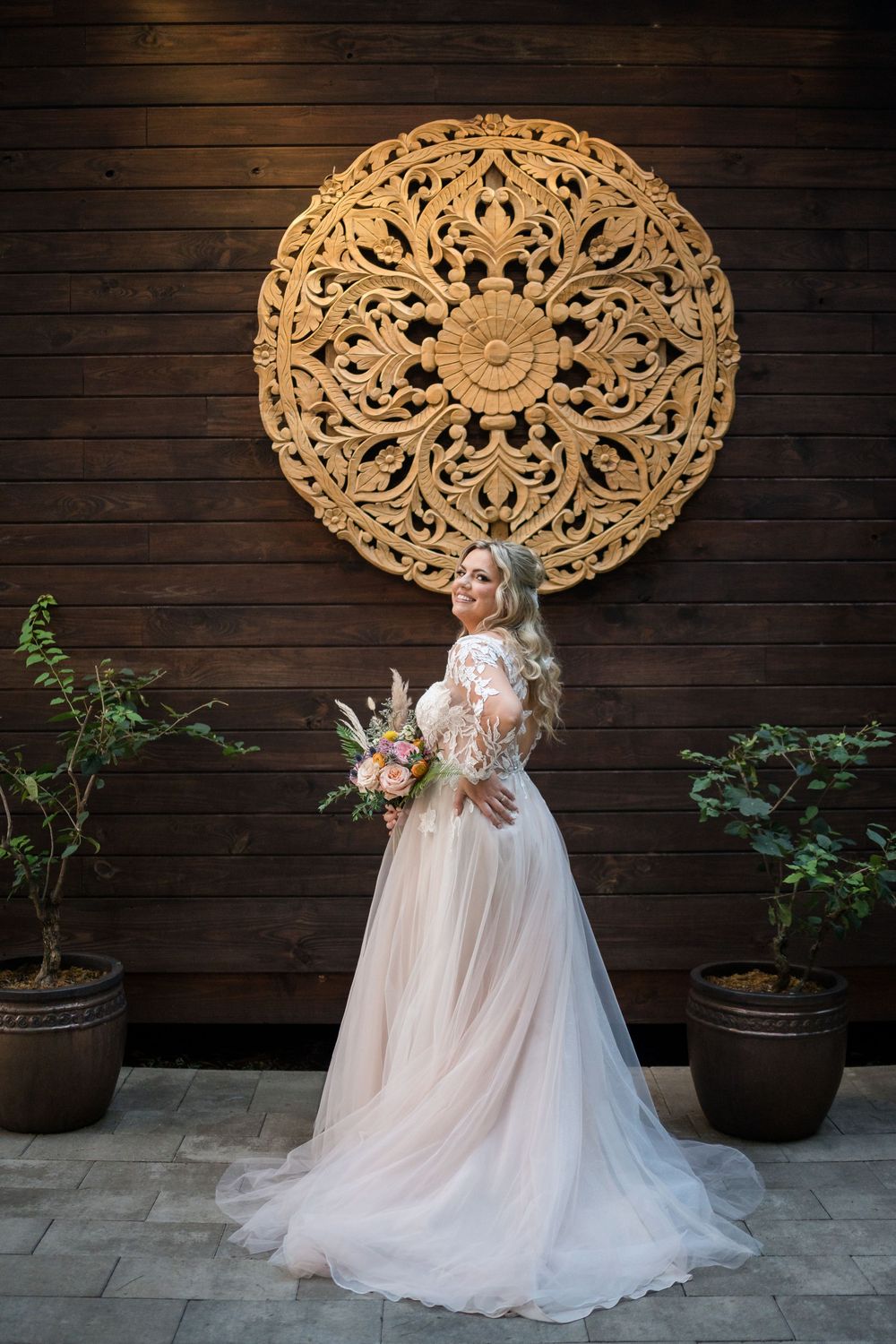 Elegant bridal portrait in front of large decorative wooden mandala wall art with potted plants on stone patio.