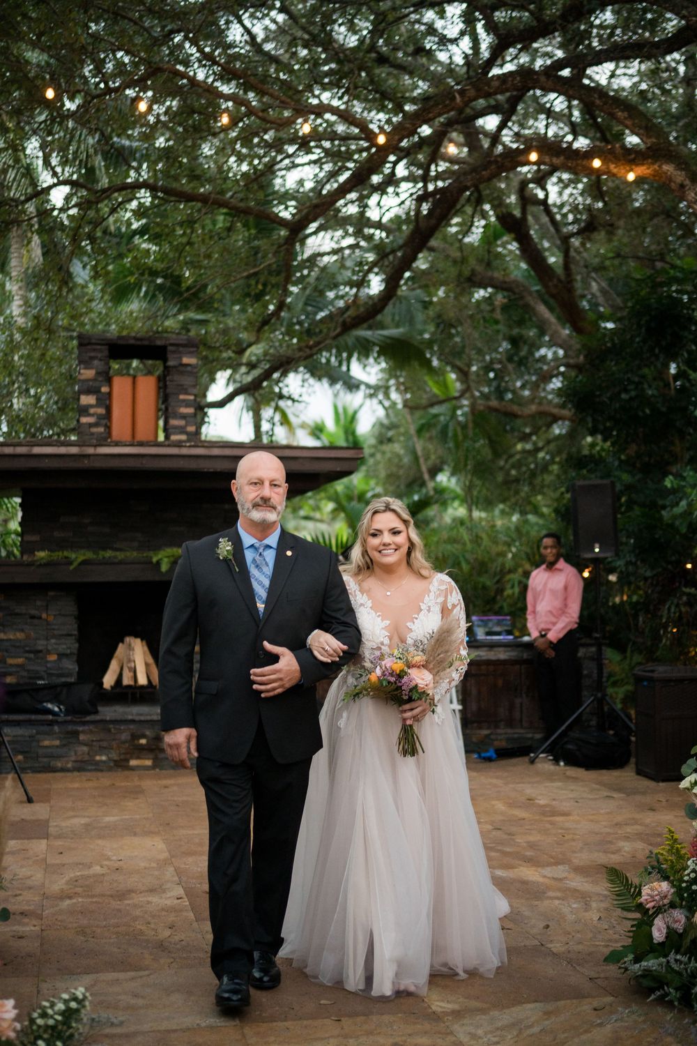 A wedding ceremony takes place on a stone patio with greenery and trees in the background.
