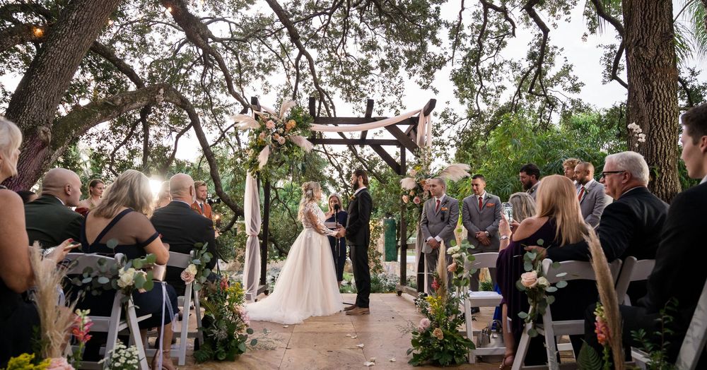 Outdoor wedding ceremony under tall trees with wooden archway and decorated aisle lined by white chairs and guests.