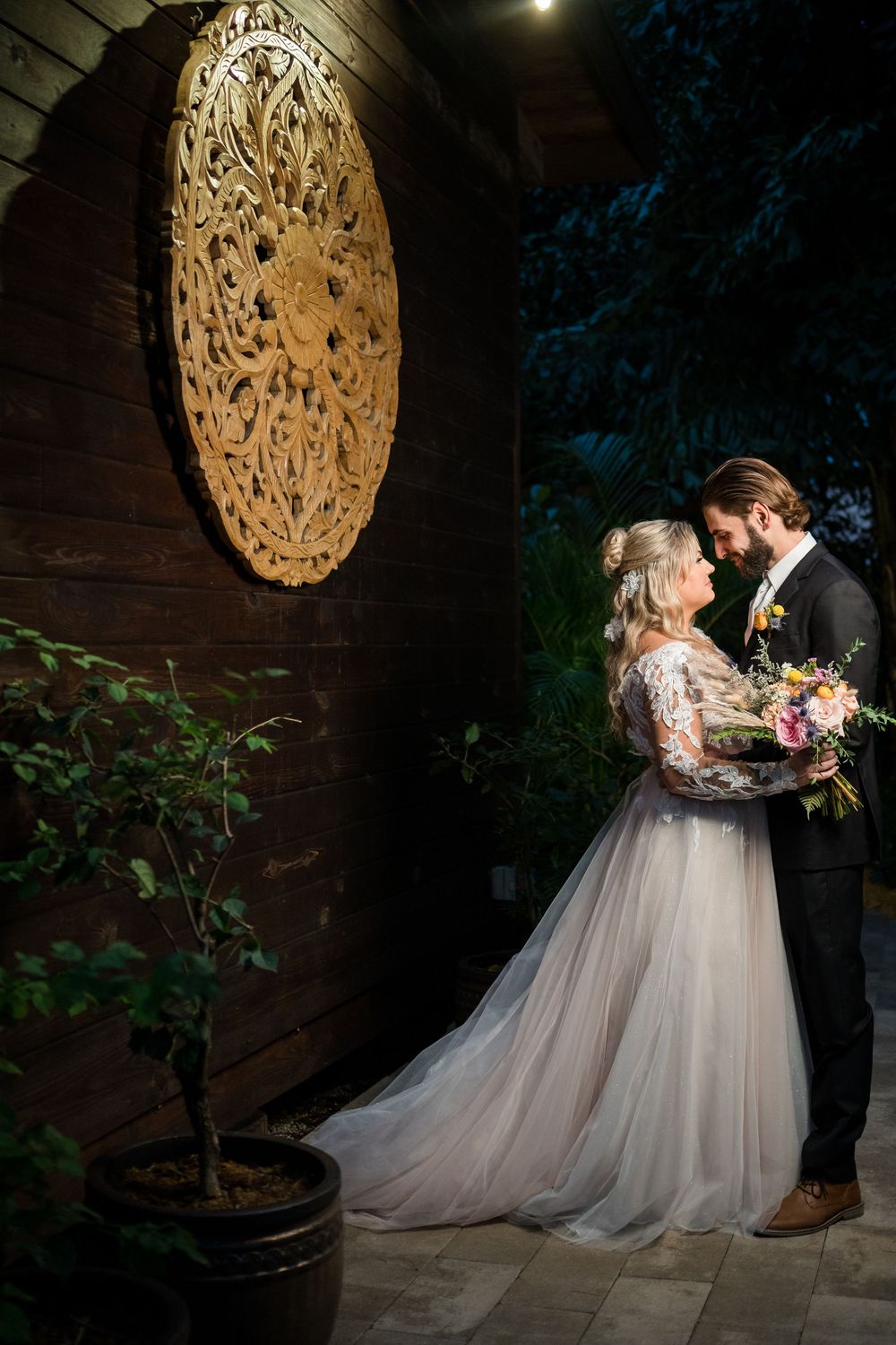 A newlywed couple shares an intimate moment under a decorative wooden mandala wall art in a garden setting.