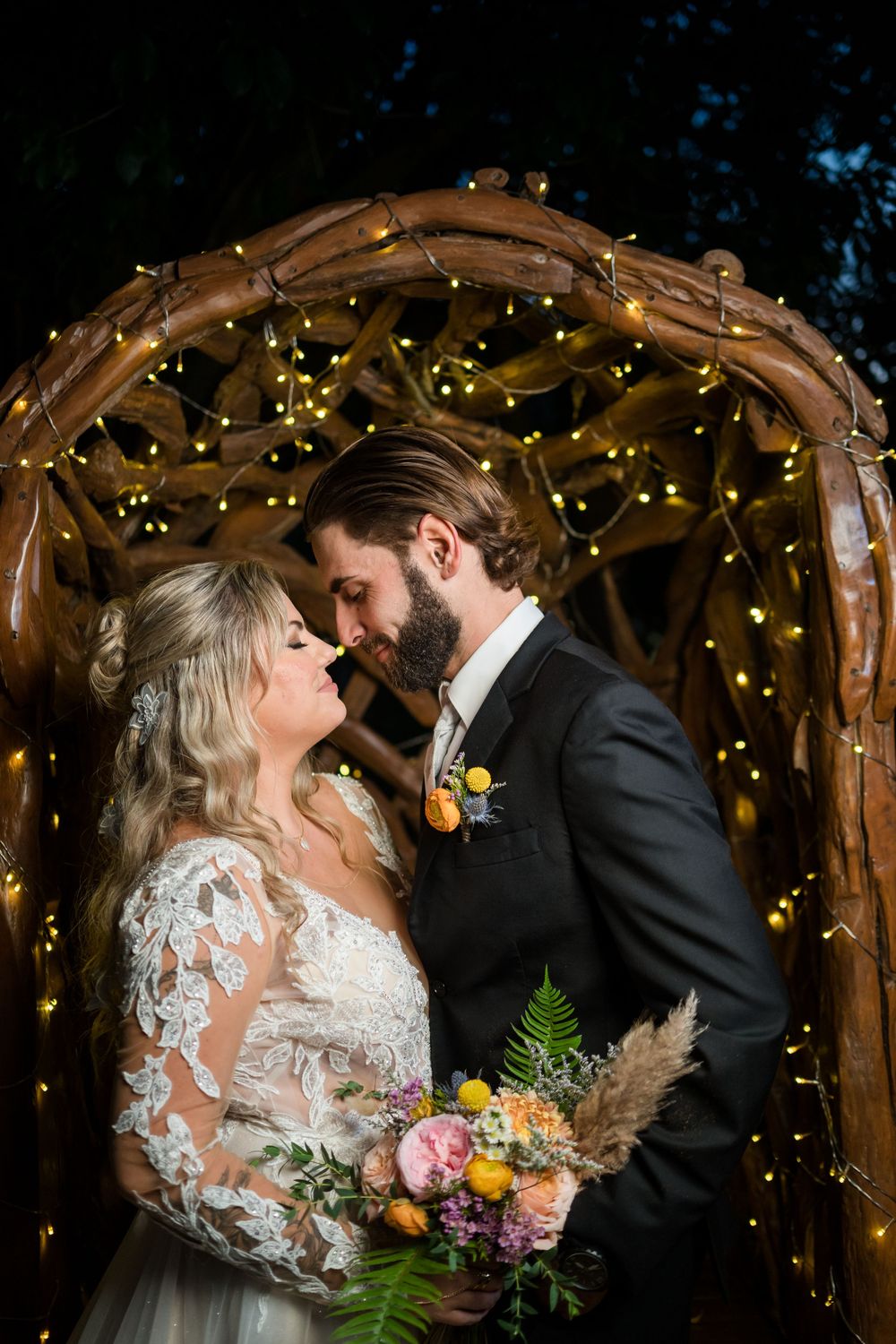Romantic wedding couple embraces under rustic wooden arch decorated with twinkling fairy lights at dusk.
