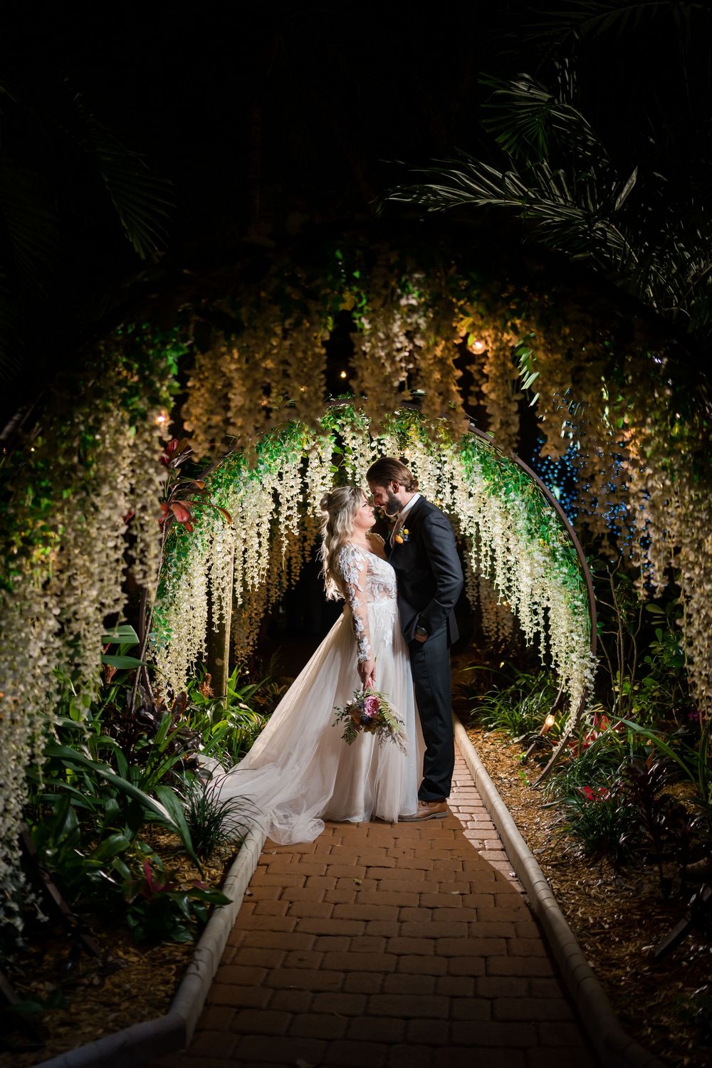 Couple stands under an illuminated floral arch tunnel draped with hanging white flowers at night.