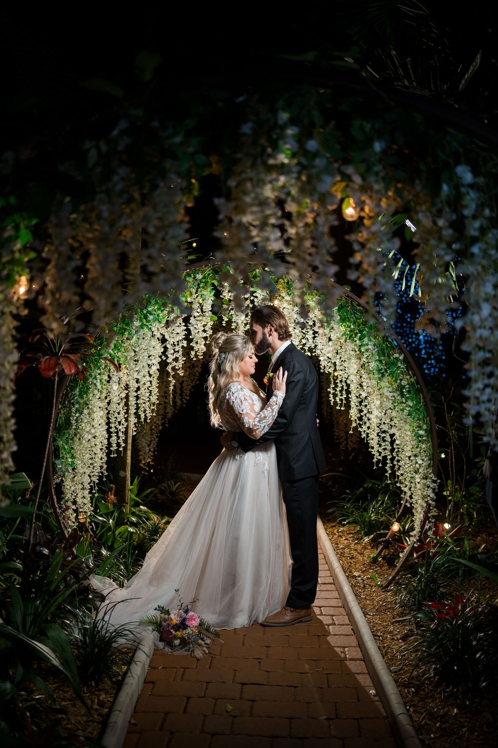 Wedding couple stands beneath enchanting canopy of cascading wisteria flowers and twinkling lights.