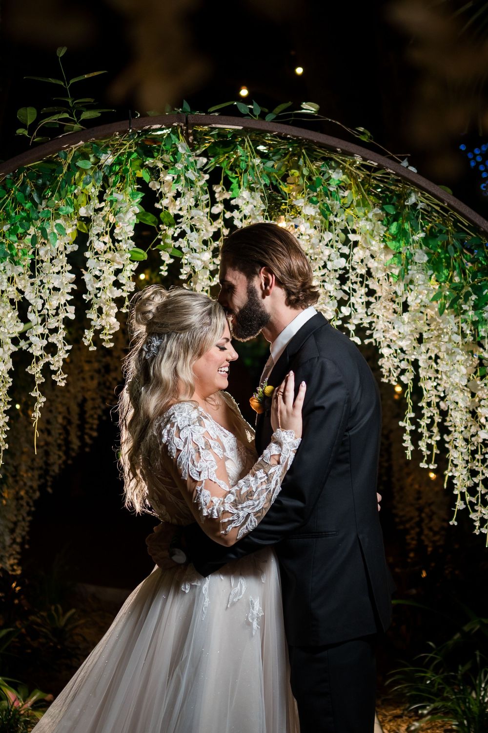 A romantic wedding couple embraces under a floral arch with cascading white wisteria and greenery at nighttime.