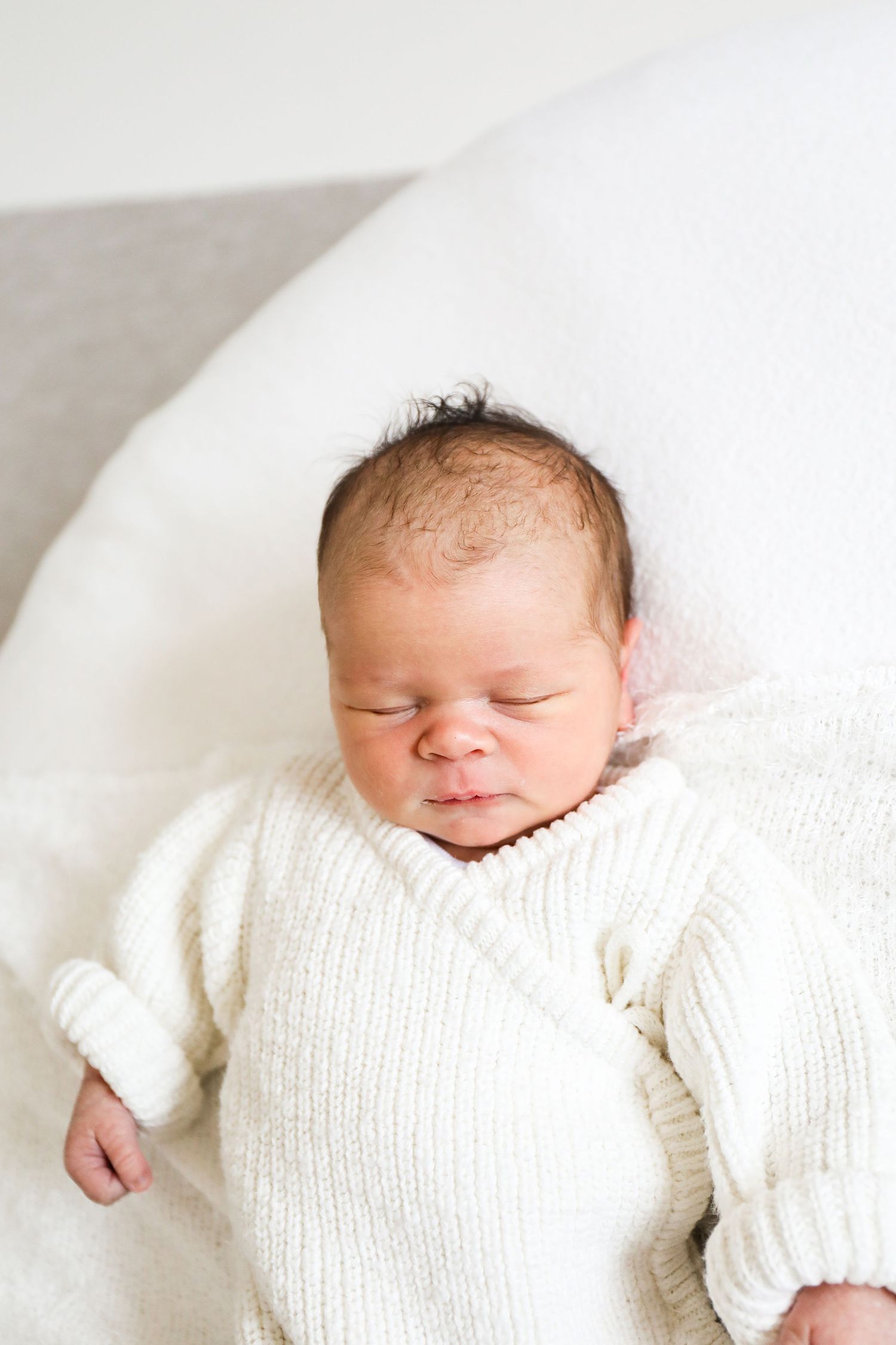 A peaceful sleeping newborn baby lies wrapped in a cozy white knit romper on soft white bedding, during newborn shoot in Nottingham