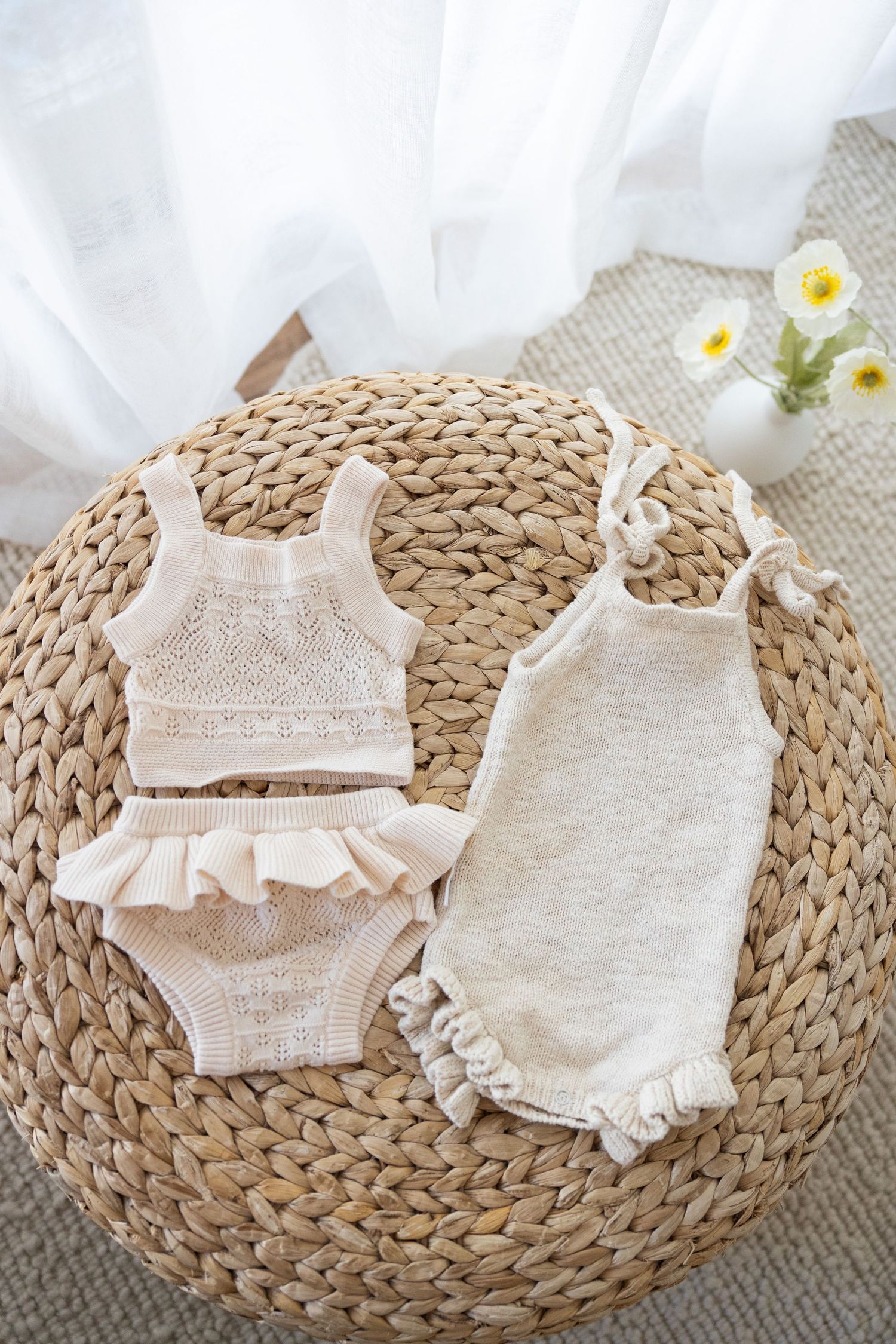 Two organic cotton baby outfits, a ruffled two-piece set and a romper, displayed on a woven wicker mat with white flowers.