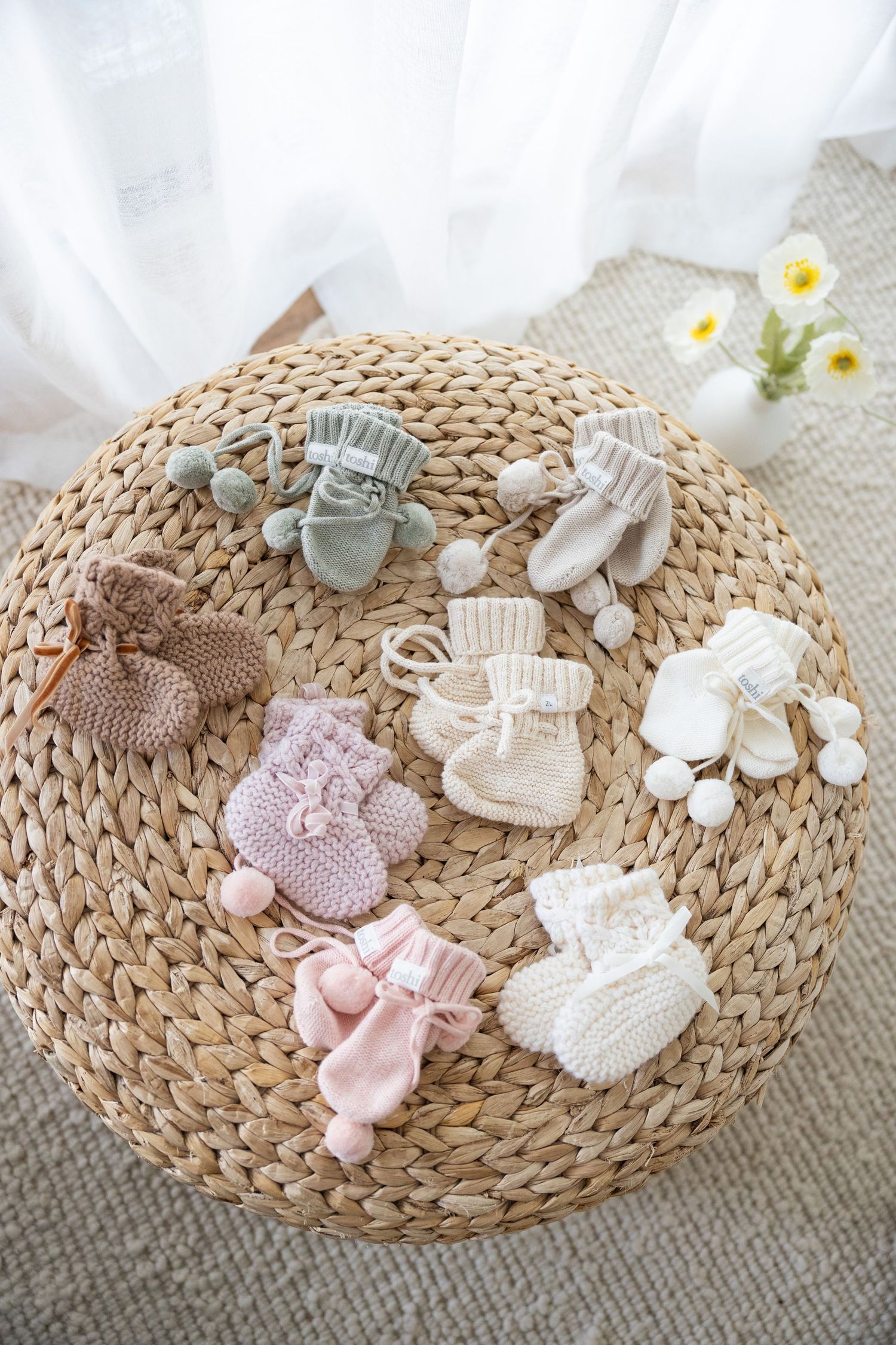 Multiple pairs of knitted baby booties in various pastel colors arranged on a round woven placemat with small white flowers.