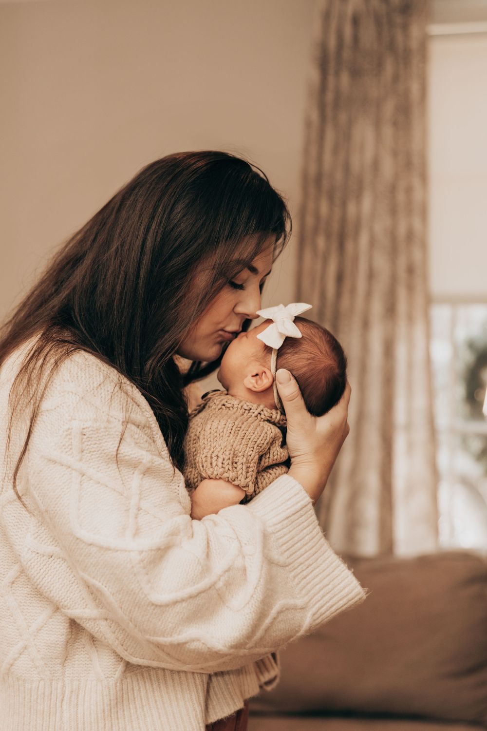 A tender moment between parent and baby in matching cream sweaters as they share a gentle embrace by a window.