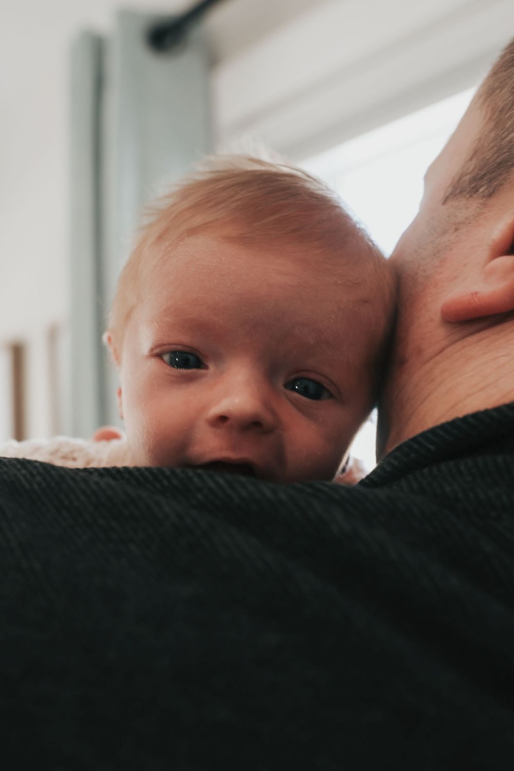A smiling baby with blue eyes rests on an adult's shoulder, looking at the camera in a bright indoor setting.