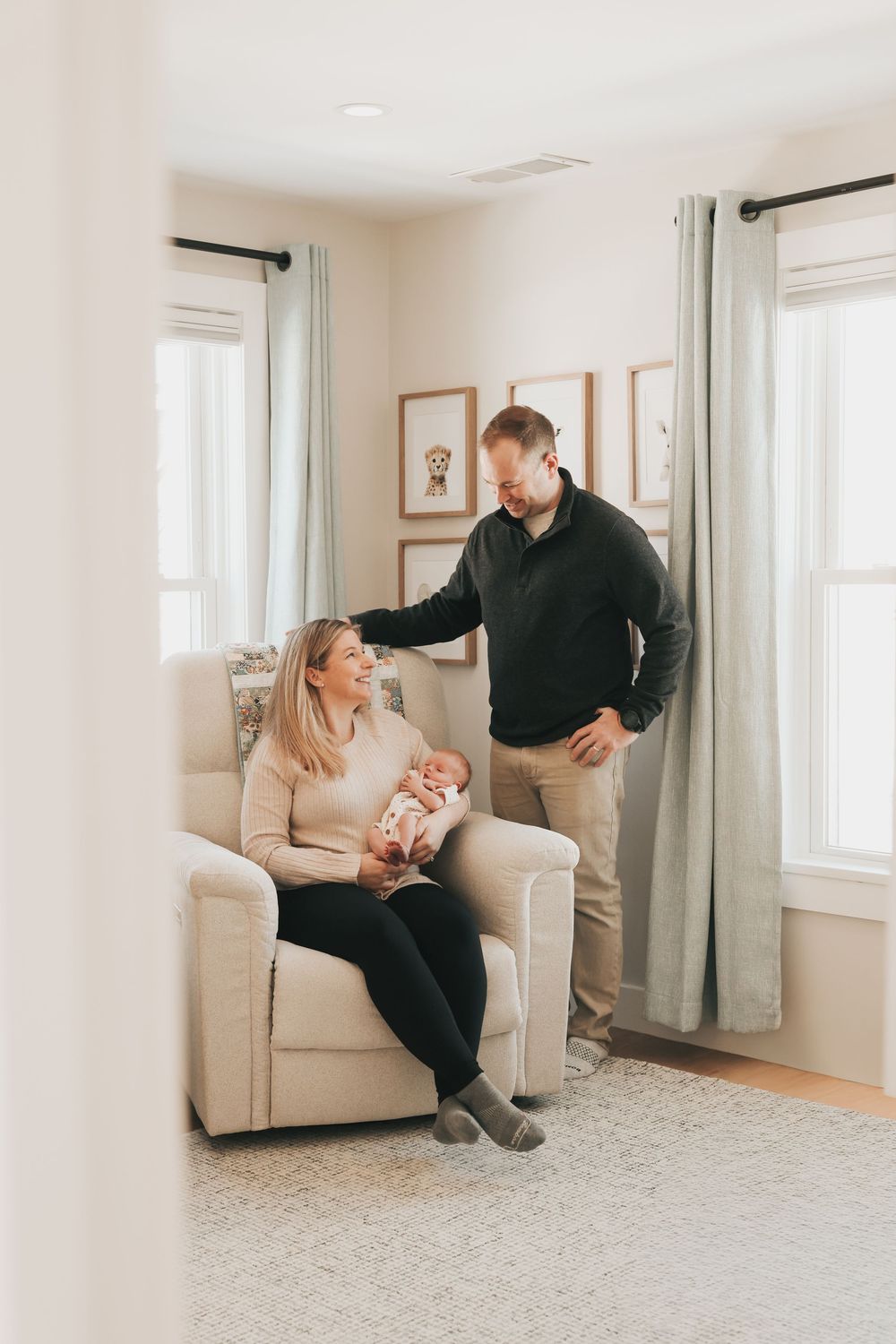 A couple smiles in a bright nursery, mother seated holding newborn baby while father stands beside her lovingly.