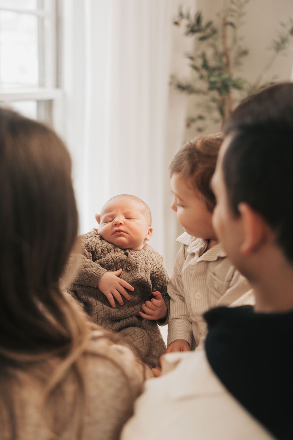 Family gathered around a sleeping newborn baby in a knit brown outfit, with a toddler sibling looking on lovingly.