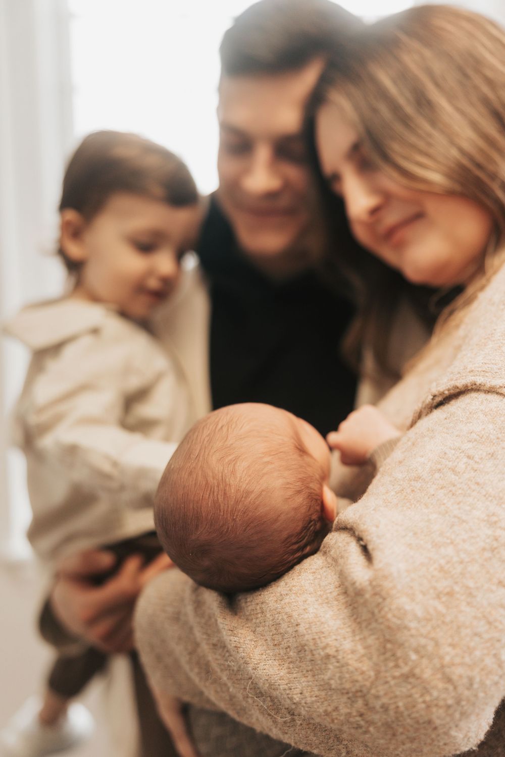 A couple smiles at their newborn baby while holding their toddler, capturing a tender family moment indoors.