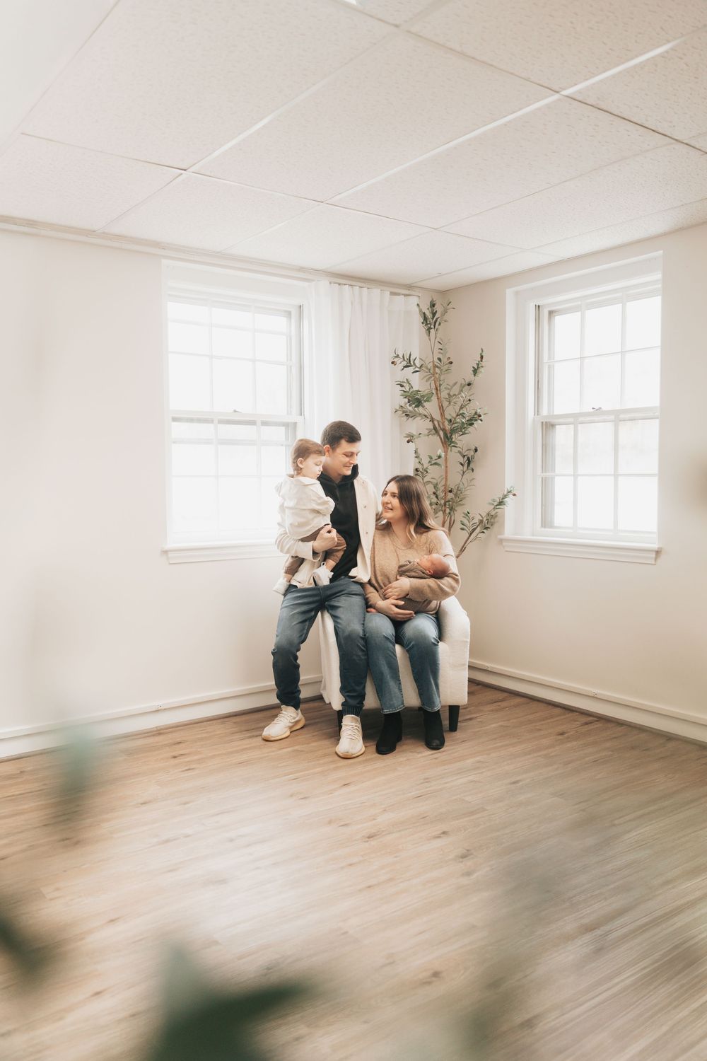 A young family with a toddler and newborn poses in a bright, minimalist room with hardwood floors and white walls.
