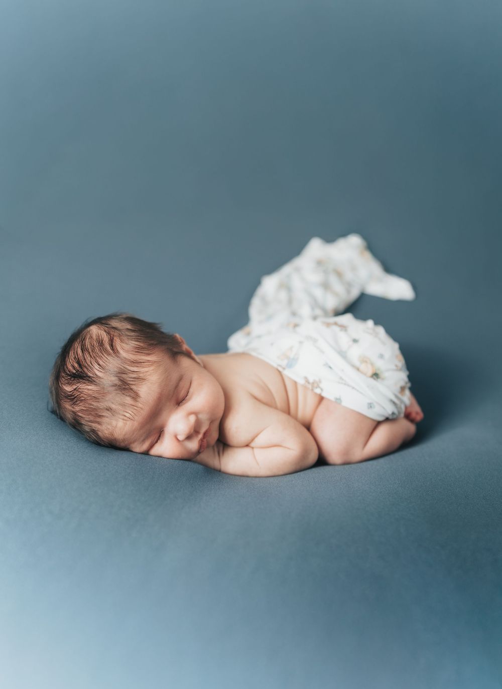 Peaceful newborn baby sleeping curled up on a soft blue backdrop, wrapped in a delicate white swaddle blanket.