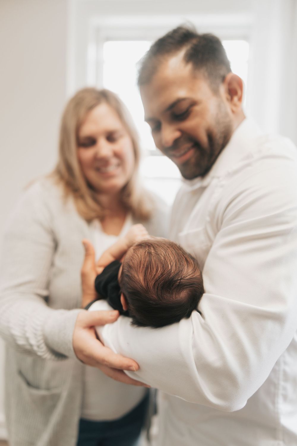 Smiling couple holding their newborn baby with dark hair in a bright room.