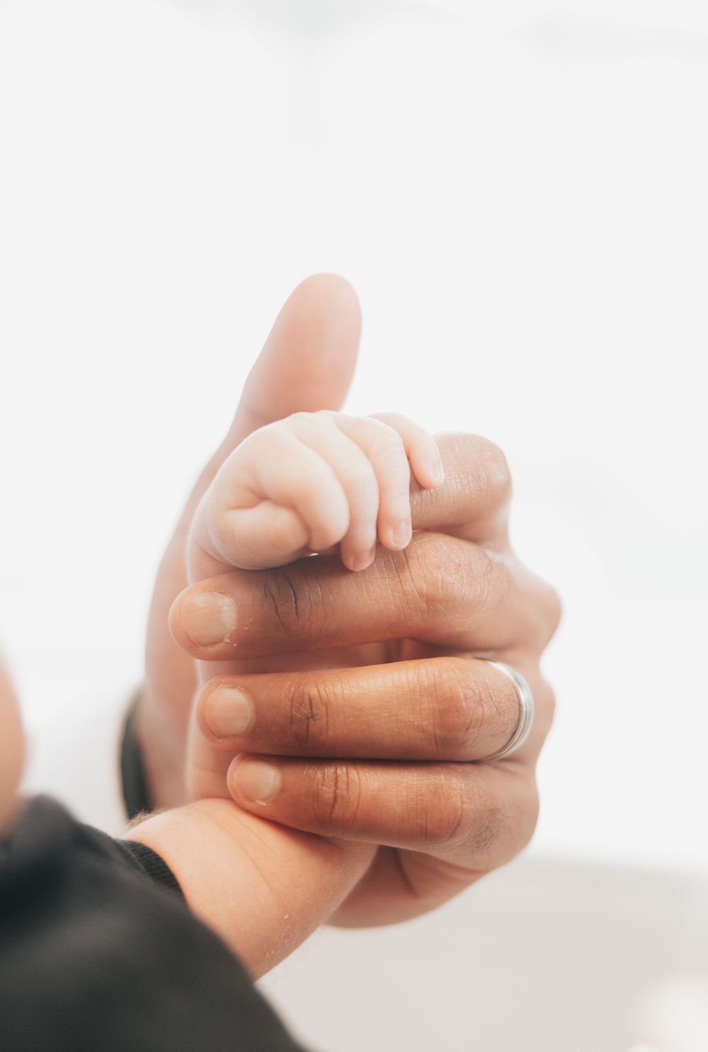 Close-up of a newborn baby's tiny hand gripping an adult's thumb, with a silver wedding ring visible.