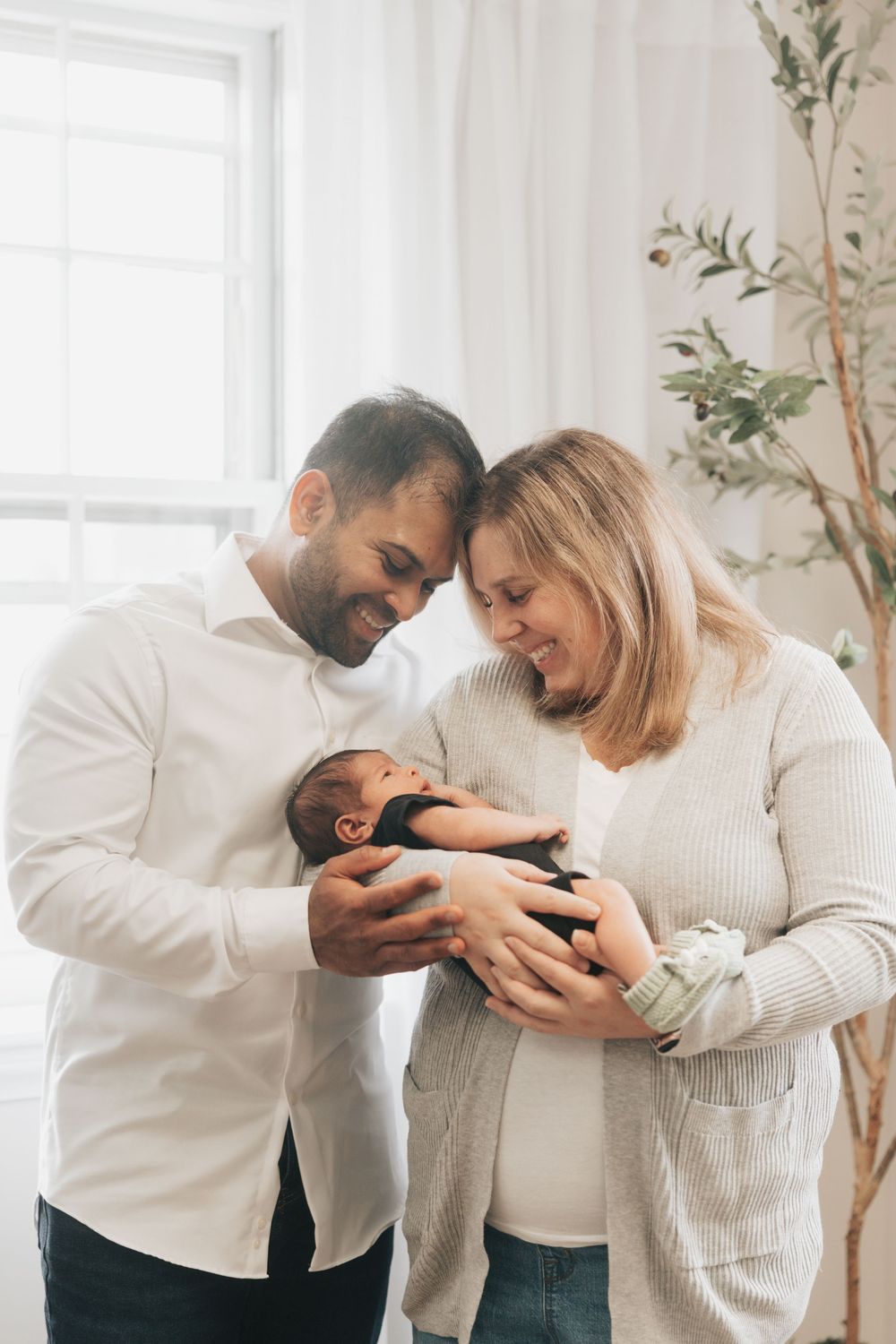 Happy parents smiling and cradling their newborn baby together by a bright window in a cozy home.