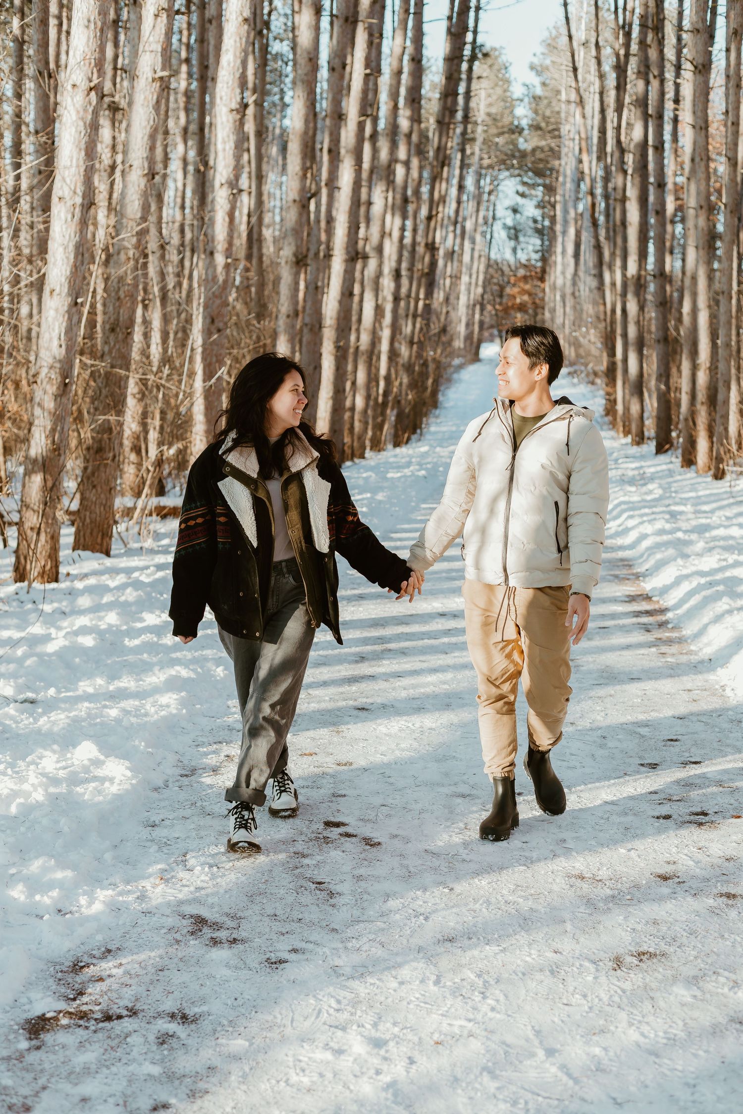 Two people walking together holding hands on a snowy trail through a winter forest landscape.