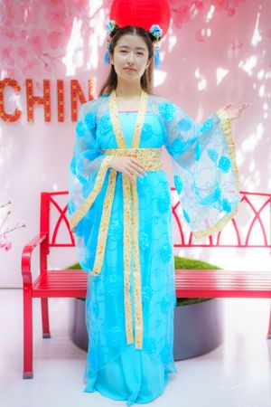 Traditional Chinese dress in bright turquoise blue with gold trim and flowing sleeves stands in front of red bench and China text.