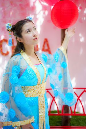 A person in a flowing blue and gold traditional Chinese hanfu dress poses with a red paper lantern against a pink backdrop.