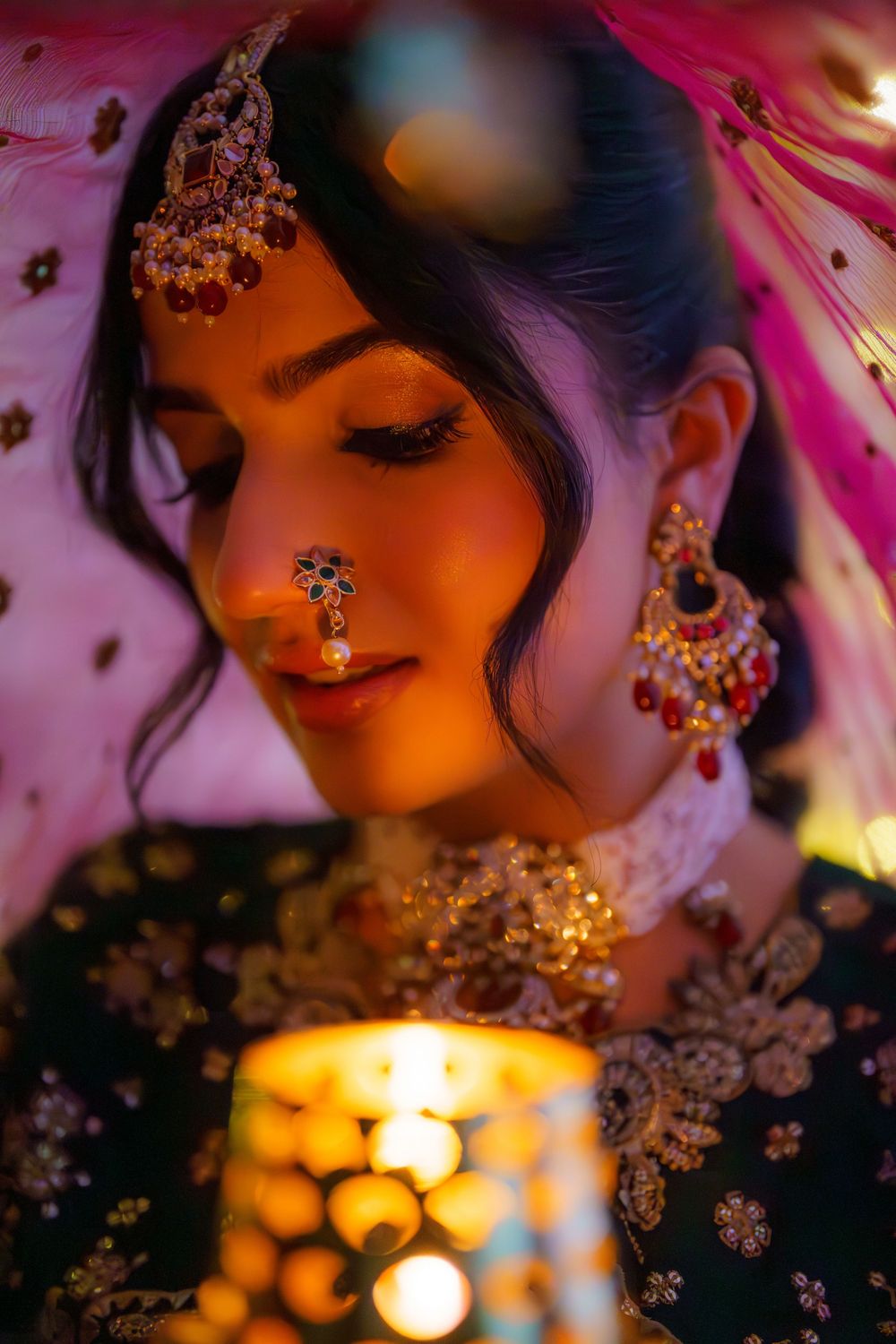 Traditional Indian bridal jewelry glimmers in warm candlelight with ornate nose ring and earrings against pink and orange tones.