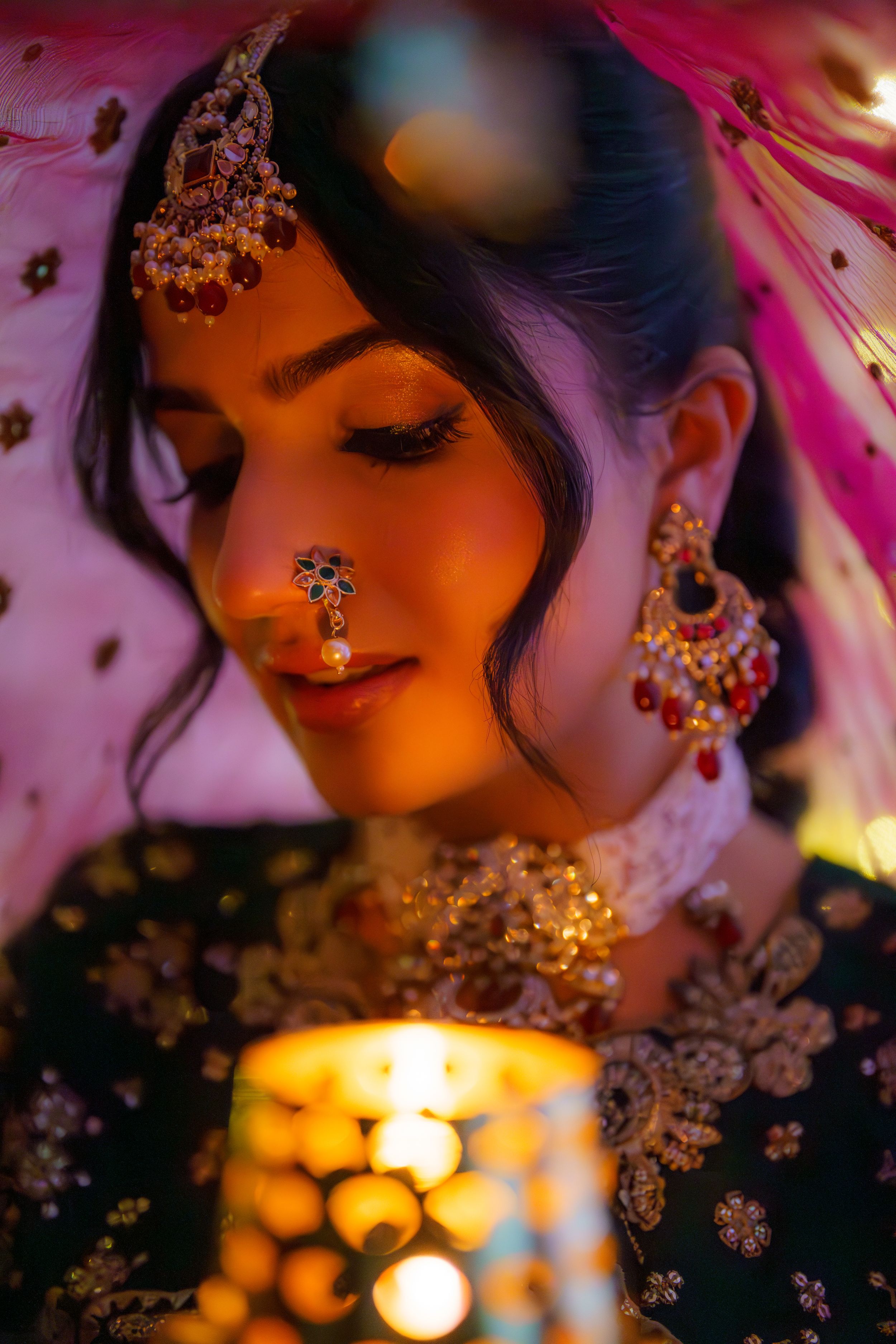 Traditional Indian bridal jewelry glimmers in warm candlelight with ornate nose ring and earrings against pink and orange tones.