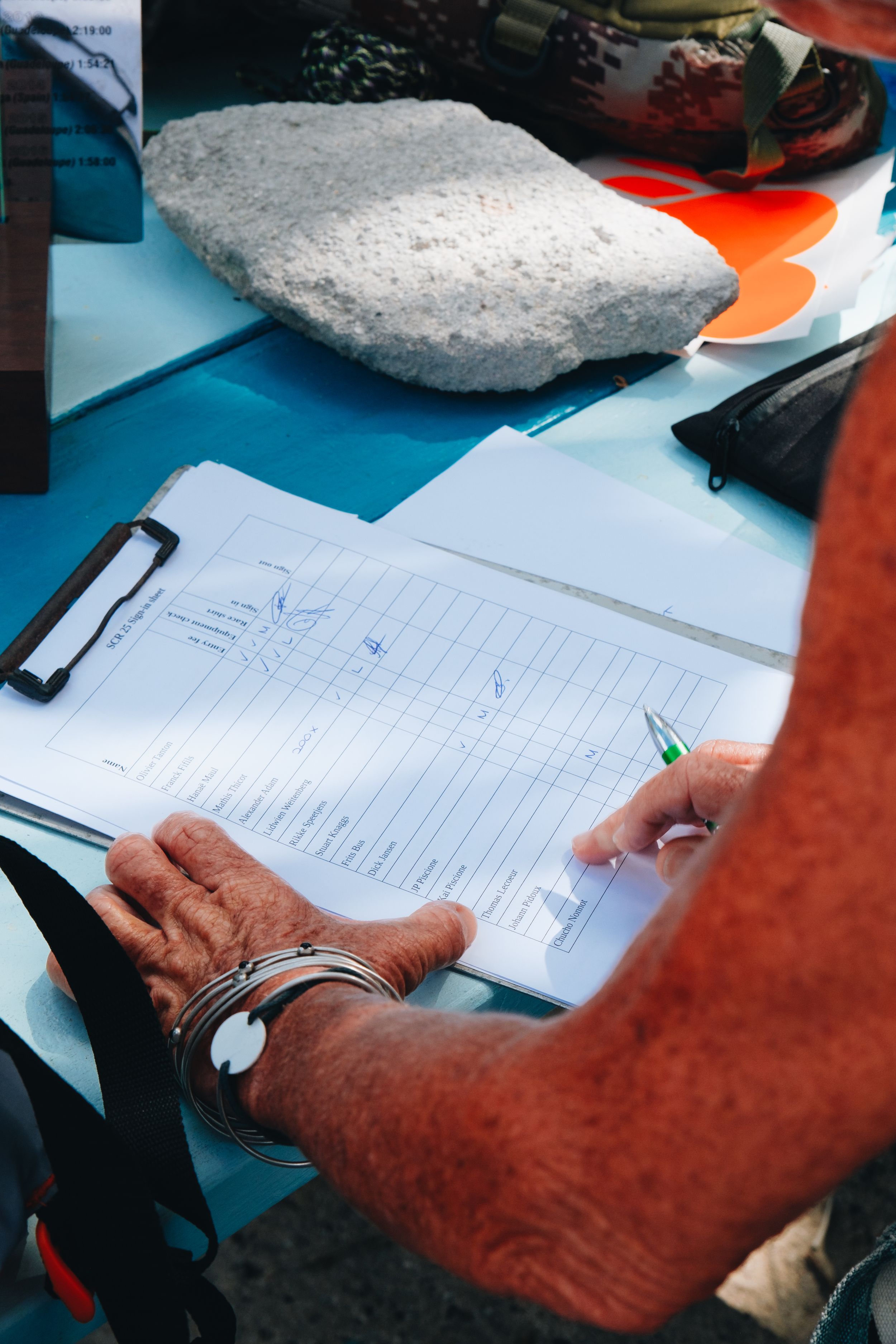 Someone wearing a watch fills out paperwork on a blue surface with a large stone nearby.