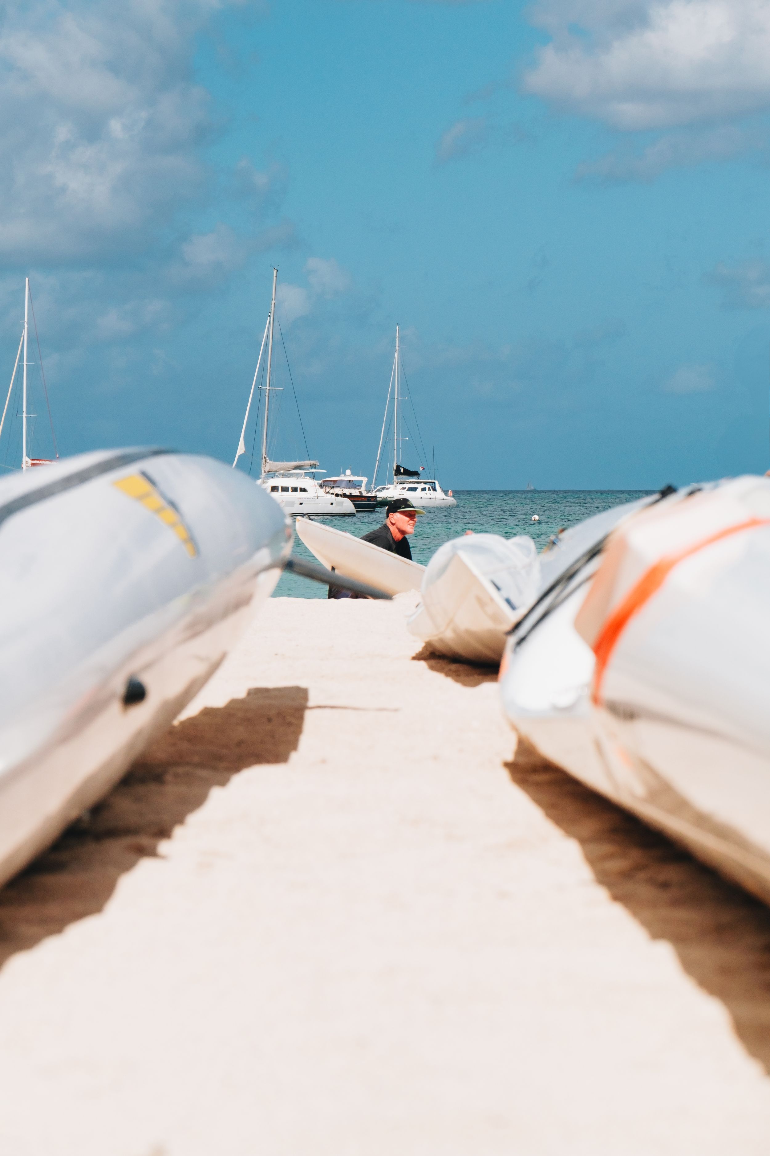 White boats and small watercraft lined up on a sandy beach with sailboats visible in the turquoise ocean background.
