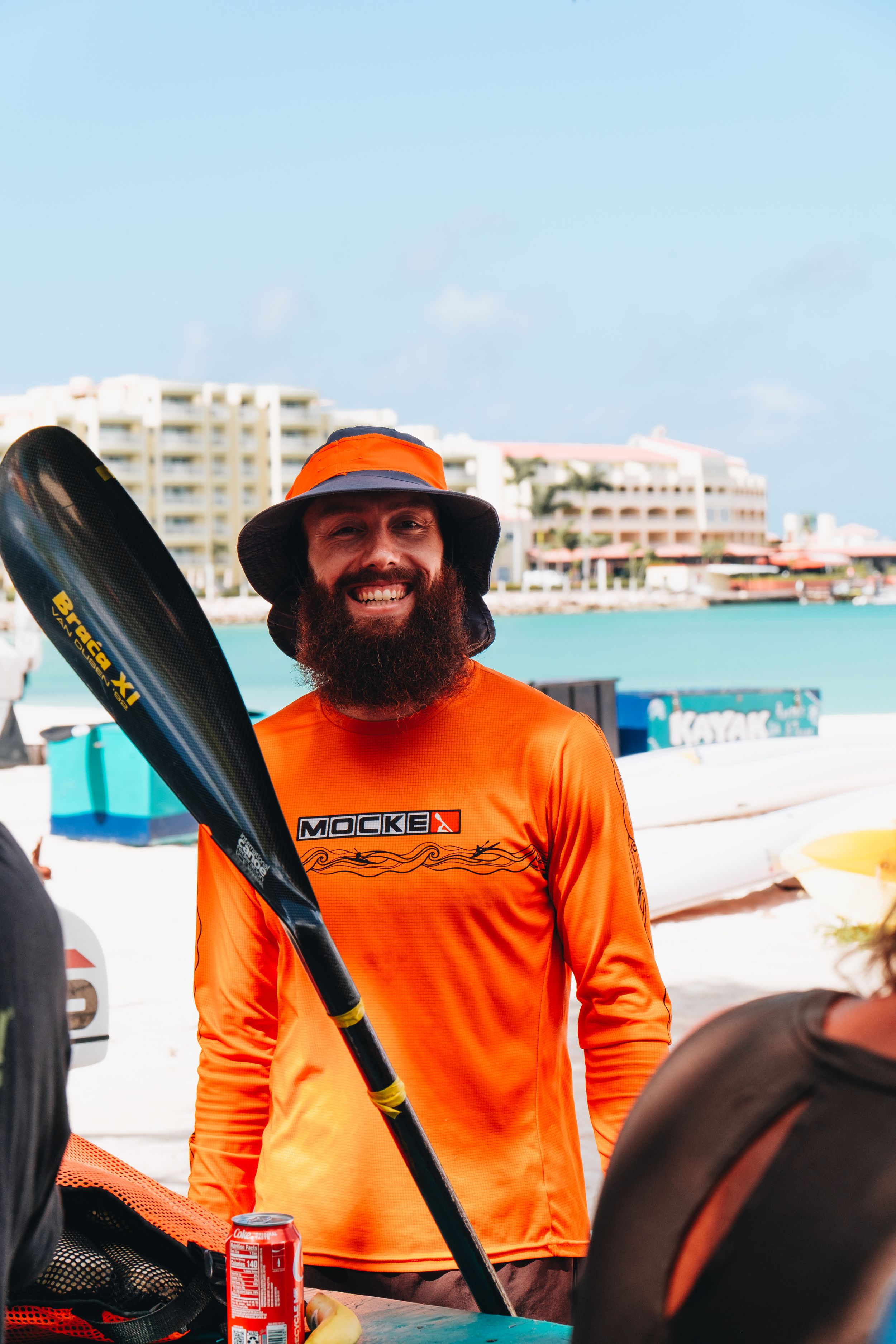 Person in bright orange sun shirt and hat holds a kayak paddle near boats and waterfront buildings on a sunny day.