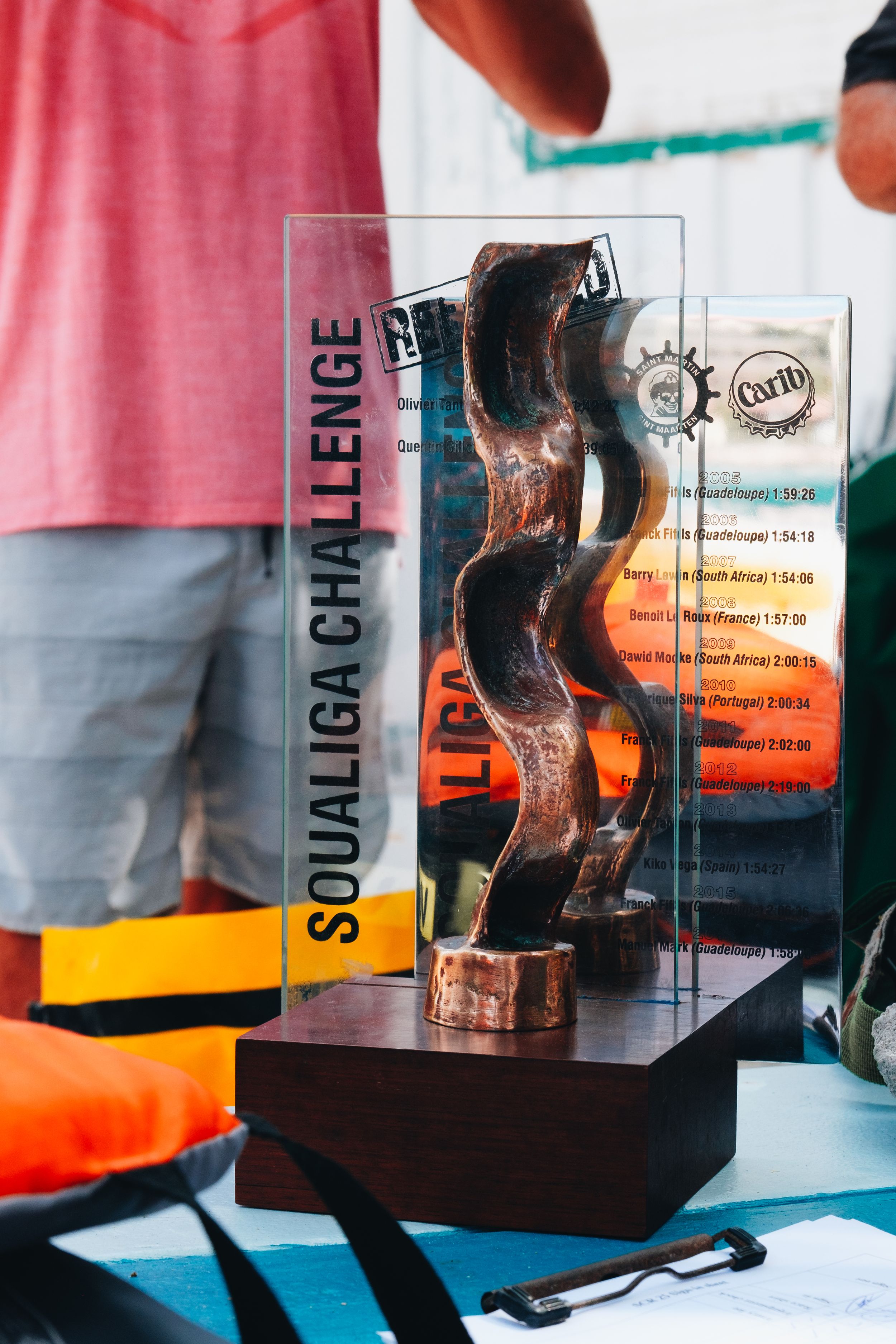 A small glass trophy or award with a curved sculpture design sits in a clear display case on a table.