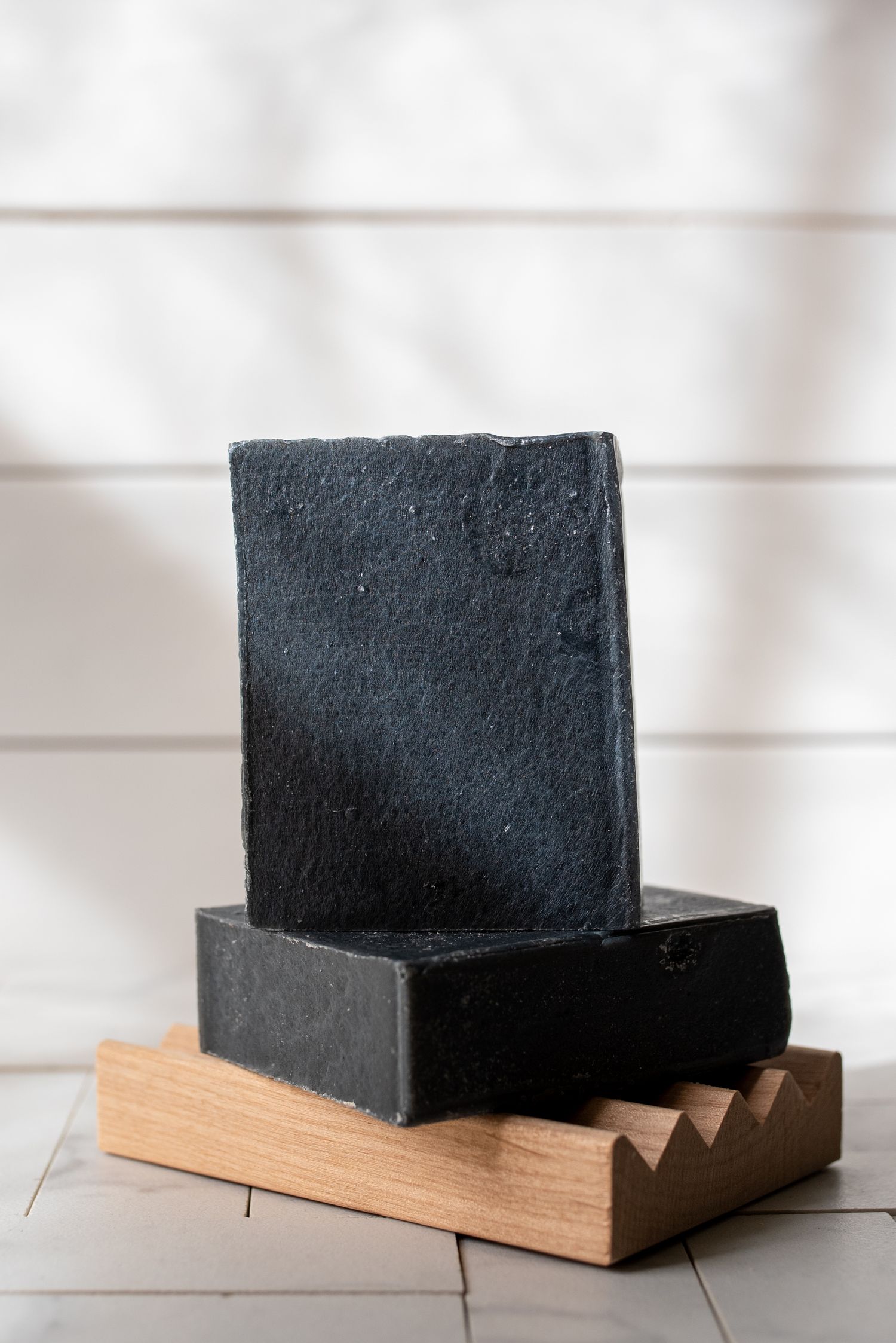Black charcoal soap bar displayed on wooden soap dish against white background.