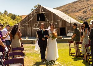 Outdoor rustic barn wedding ceremony with guests seated on purple chairs.