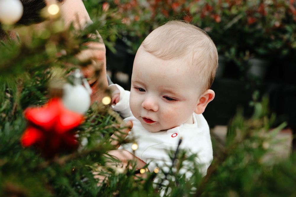 Baby reaches toward Christmas tree ornament while wearing white holiday outfit.
