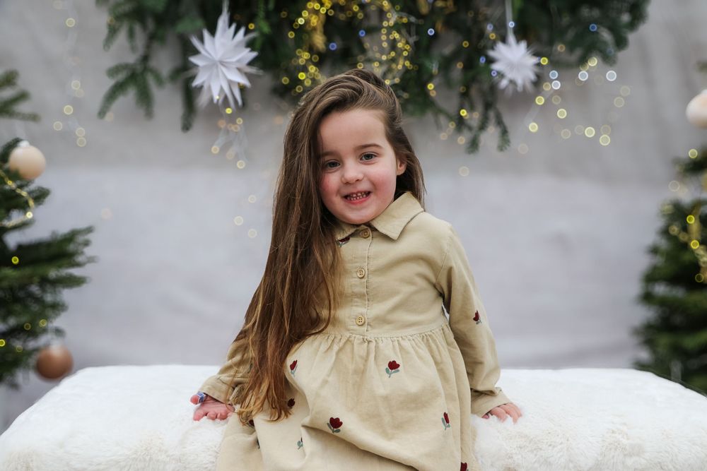 Child in beige dress poses on white bench with holiday decorations and snowflakes.