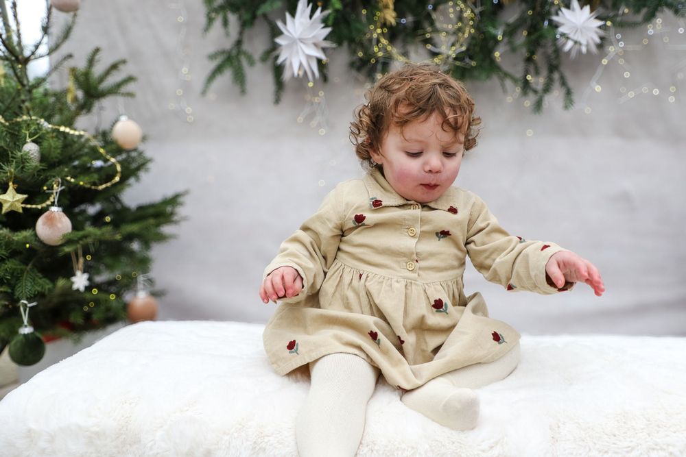 Baby in beige dress plays joyfully in front of decorated Christmas tree with ornaments and snowflakes.