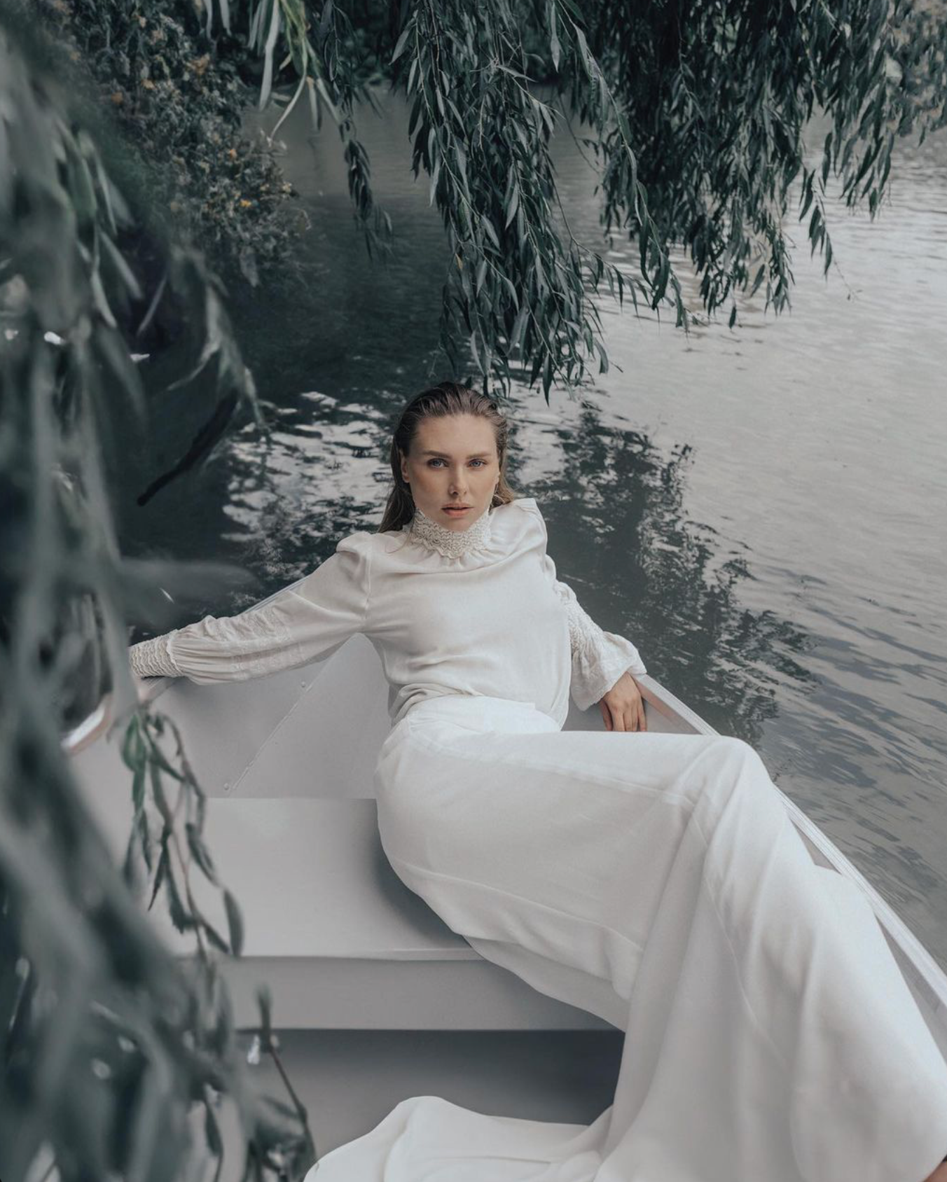 Person in white dress reclines elegantly in boat under weeping willow branches over water.