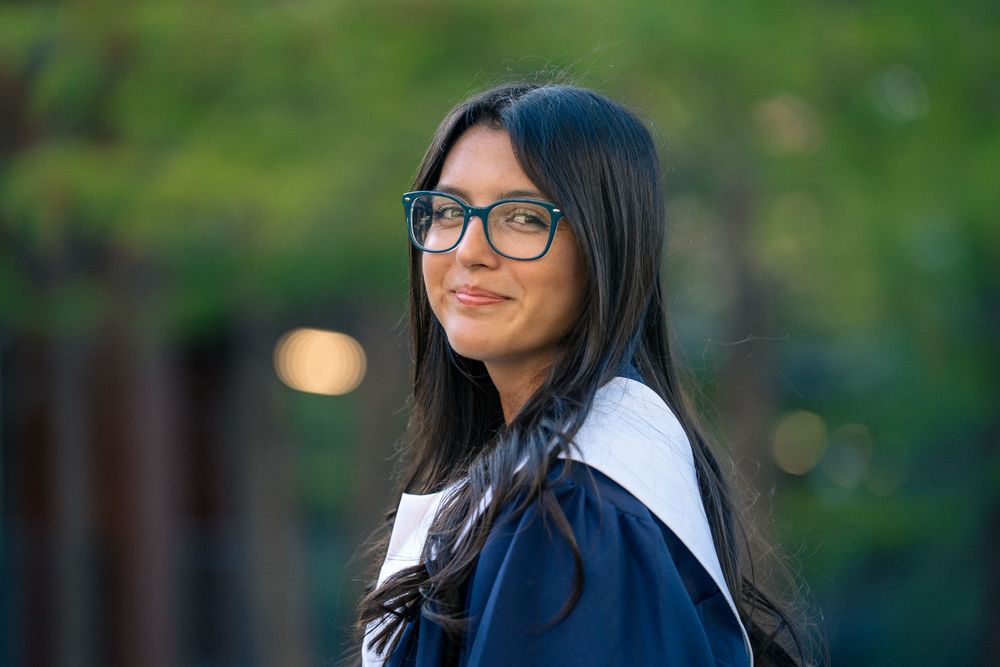 Young woman with long dark hair and glasses smiles while wearing a navy blue jacket outdoors.