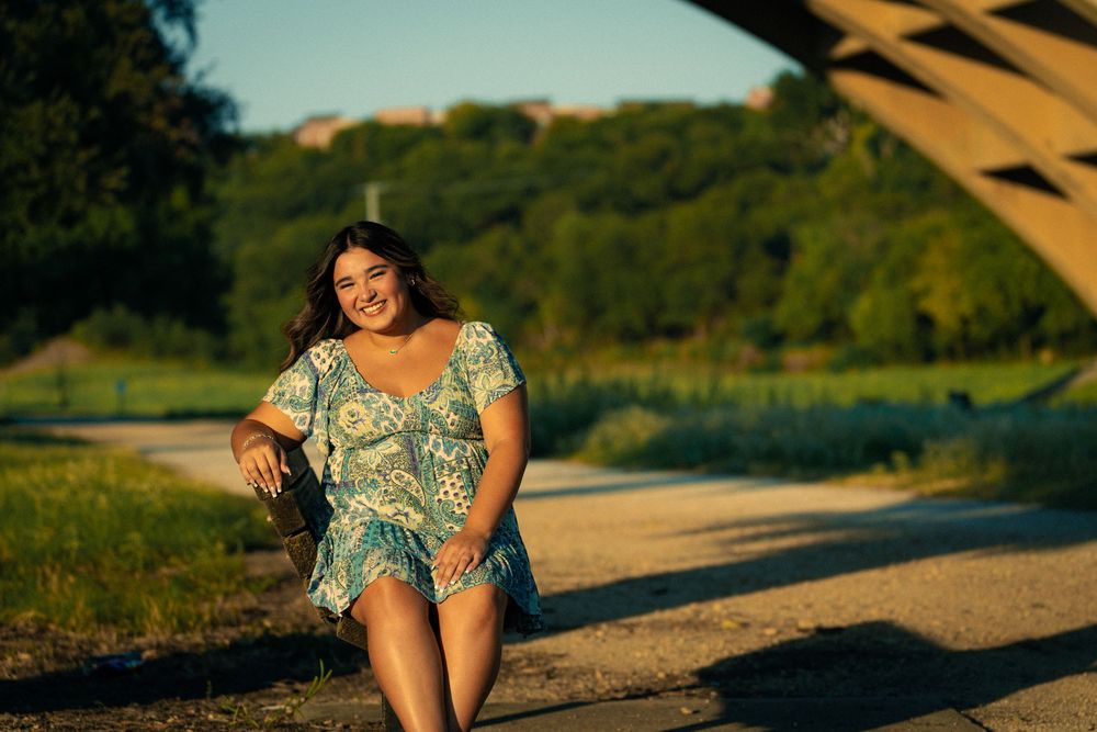 A person in a floral dress sits on a sandy path during golden hour with trees and hills in the background.