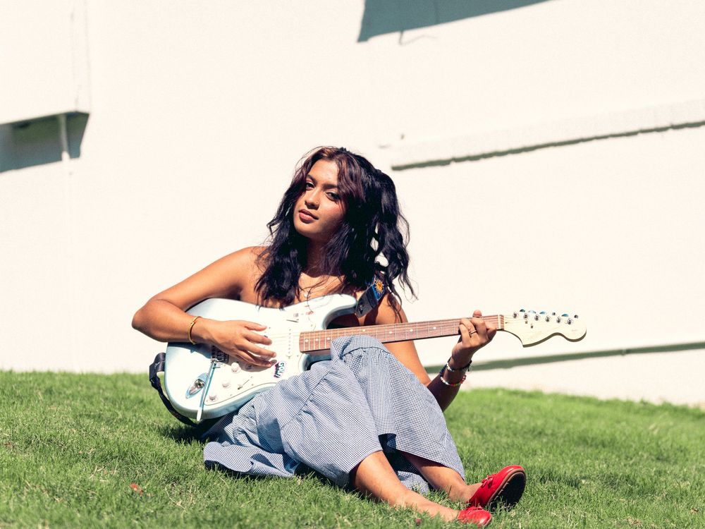 A woman in a light blue dress lies on a grassy hillside while smiling and relaxing outdoors on a sunny day.
