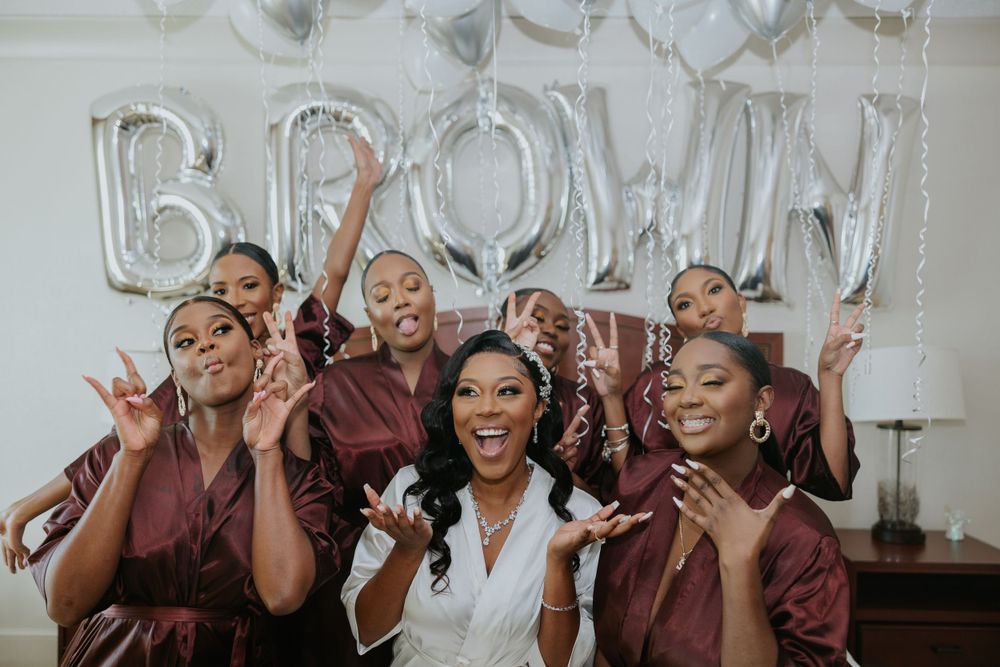 Wedding party in burgundy dresses celebrating with bride in white, with silver BRIDE balloon letters in background.