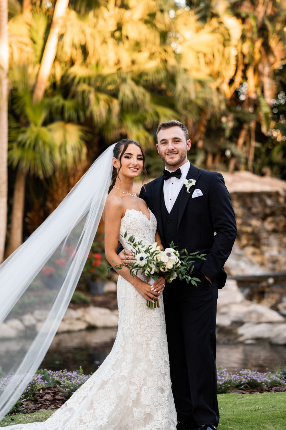 A wedding couple poses outdoors in formal attire with palm trees and a stone wall in the background at sunset.