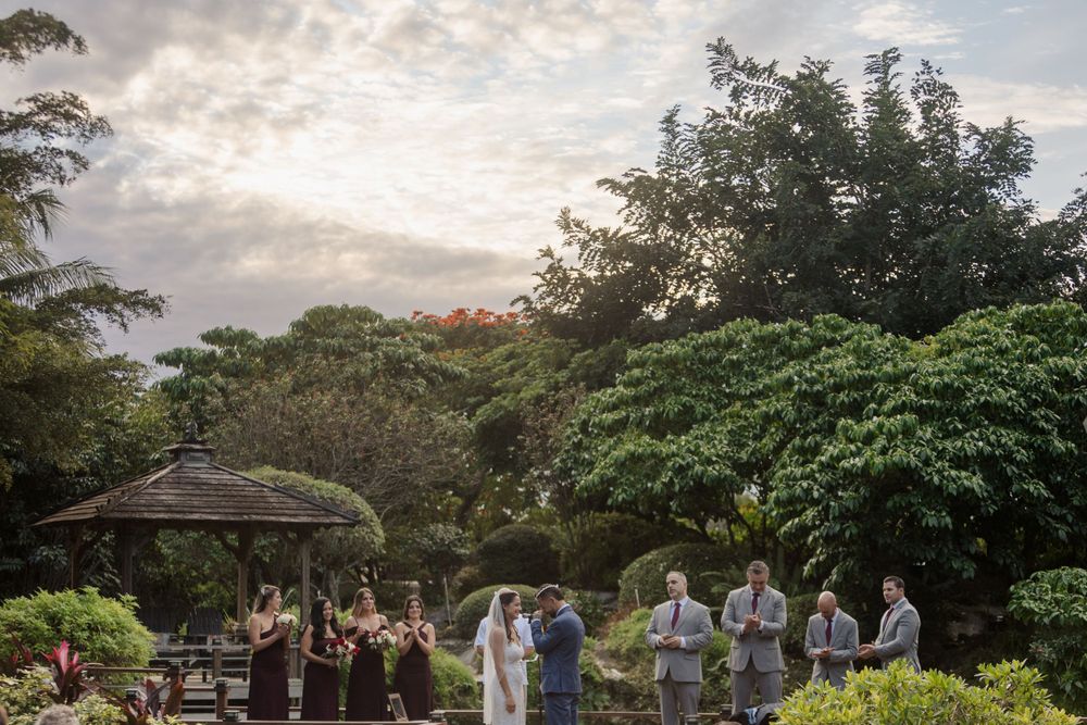 Outdoor garden party gathering at sunset with guests mingling under a gazebo among lush tropical greenery.