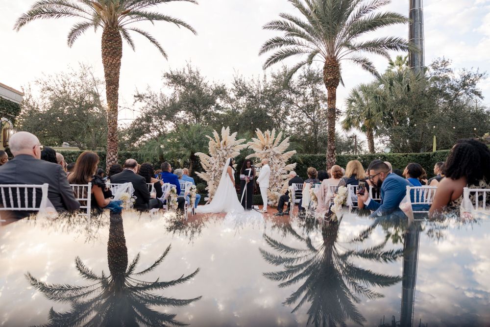 Wedding ceremony reflected in mirrored floor with tropical palm trees and guests seated outdoors in a garden setting.