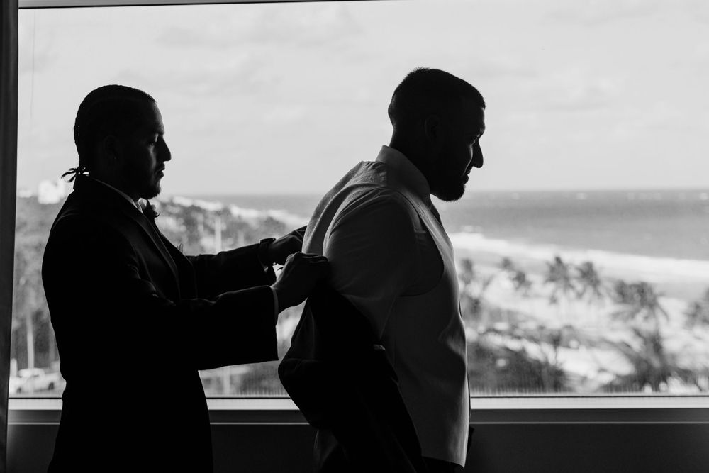 Black and white silhouette of two people standing by a balcony overlooking a tropical beach with palm trees.