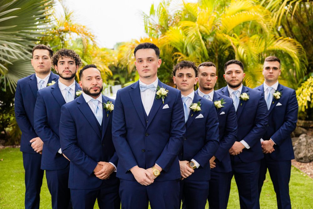 A wedding party in navy blue suits with white boutonniere flowers poses on a lush tropical garden path with palm trees.