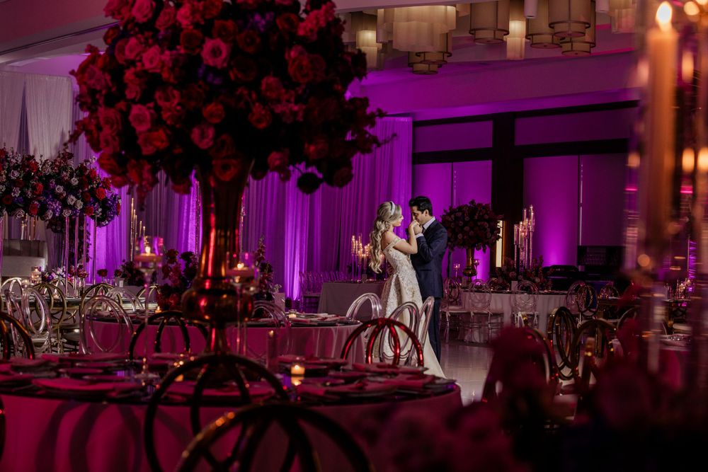Elegantly decorated wedding reception ballroom with purple uplighting, red floral centerpieces, and dancing figures on the floor.