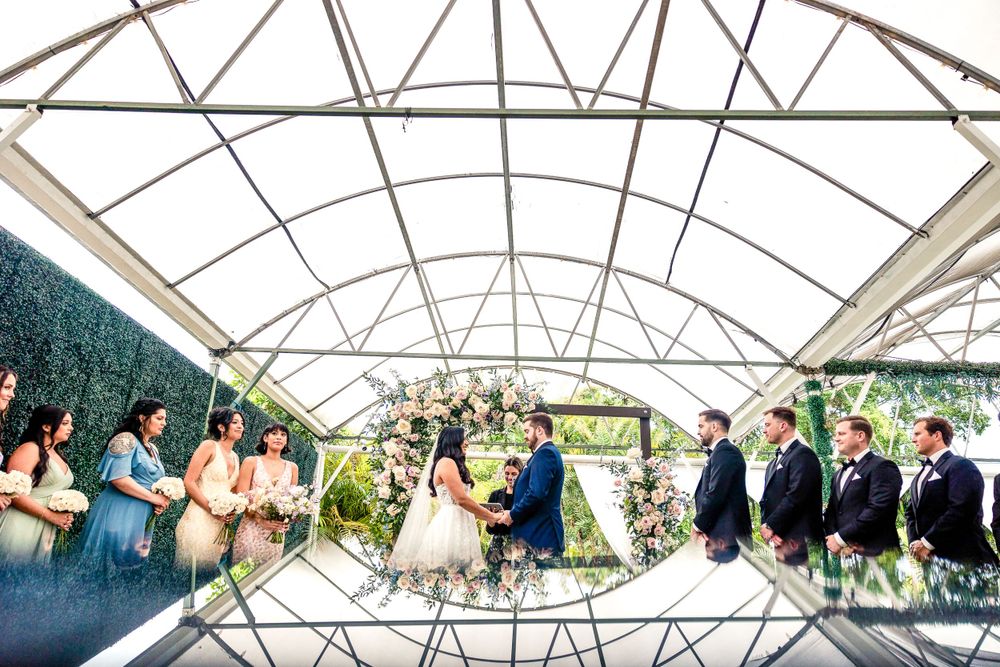 Wedding party stands beneath arched greenhouse structure during outdoor ceremony with greenery and natural light.