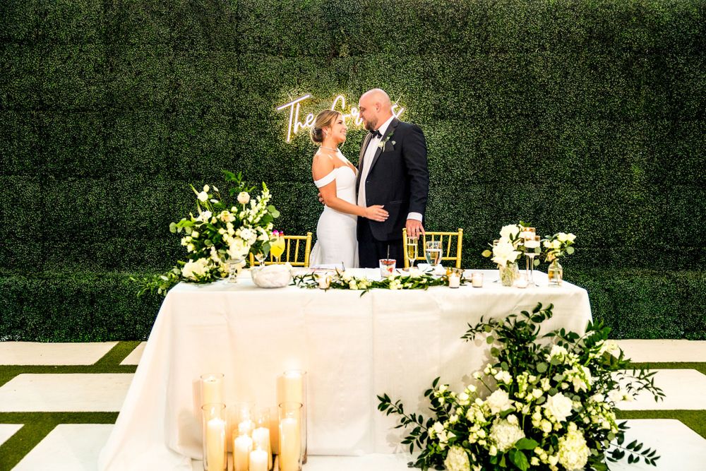 A wedding couple shares a moment at an elegant reception table with white floral arrangements and candles against a green hedge wall.