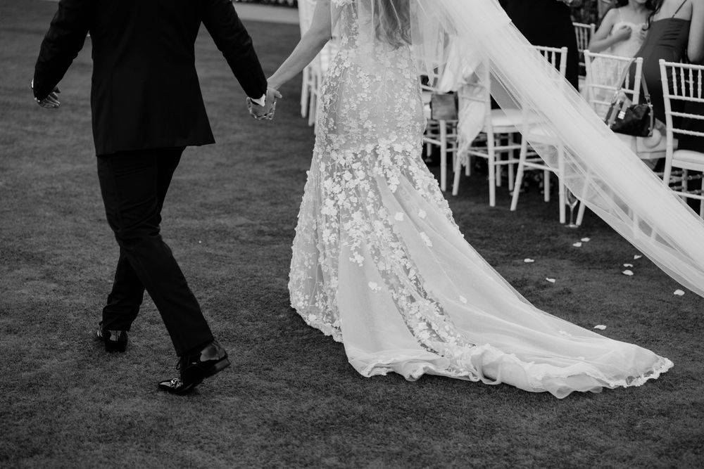 Black and white photo of wedding dress details with lace train flowing behind as couple walks together outdoors.